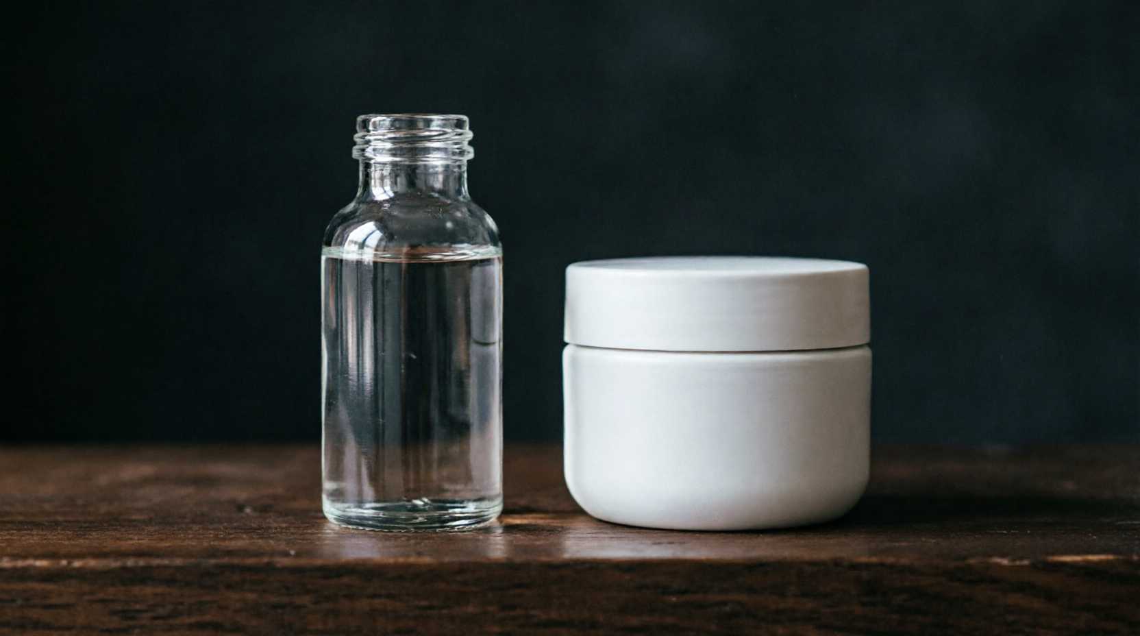 A clear glass bottle of glycerine standing next to a white cosmetic cream jar on a dark shelf.
