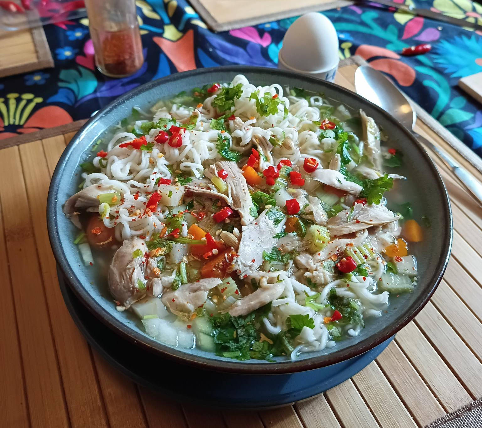 A bowl of golden-yellow chicken broth with ramen noodles, fresh cilantro, red chilies, and a perfectly soft-boiled egg.